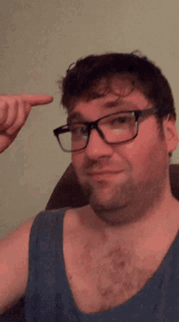 a man wearing glasses and a blue tank top points his finger at his head