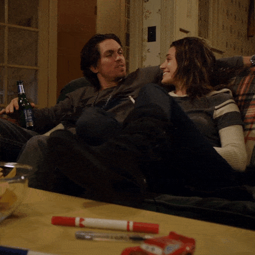 a man and woman sit on a couch with a bottle of corona