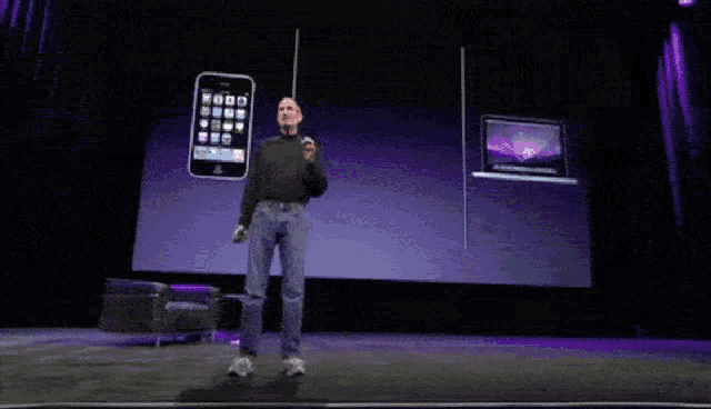 a man stands on a stage in front of a large screen with a phone and laptop on it