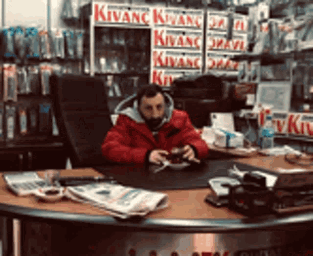 a man in a red jacket sits at a desk in front of a kivanc display