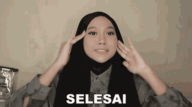 a woman wearing a hijab is making a selesai sign