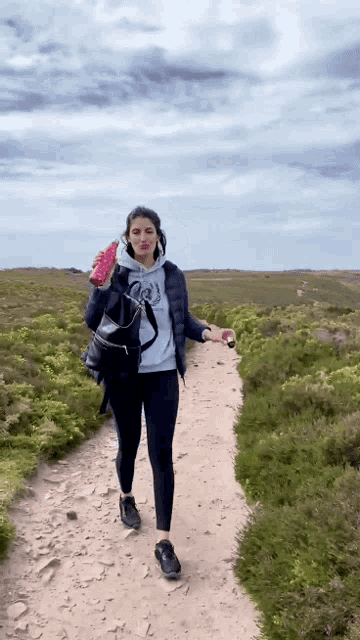 a woman is walking down a dirt path holding a pink bottle
