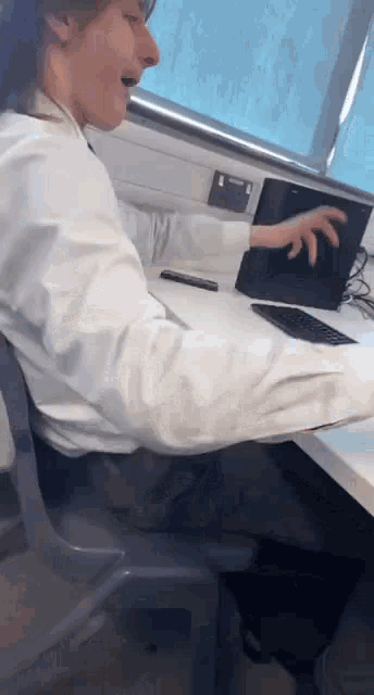 a man in a white shirt is sitting at a desk using a laptop .