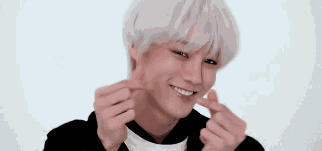 a young man with white hair is smiling and making a heart with his hands .