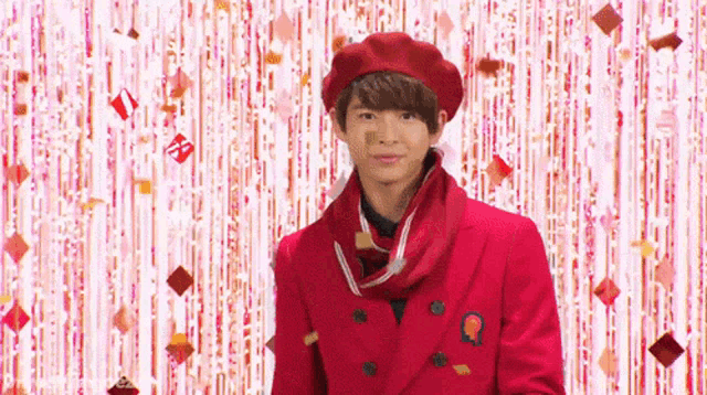a young man in a red suit and beret is standing in front of a pink curtain with confetti falling around him .