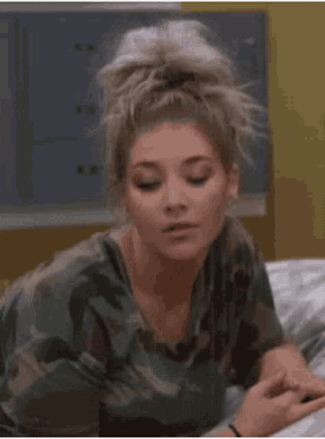 a woman in a camouflage shirt is sitting on a bed and looking at her phone .