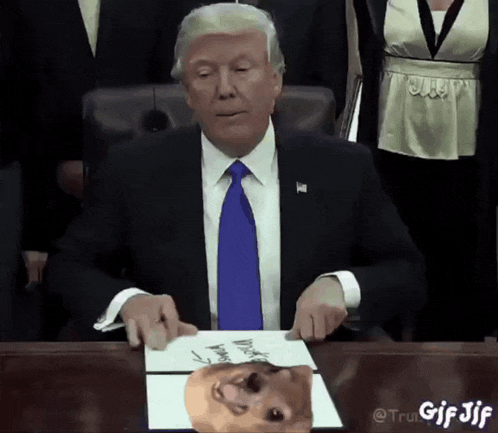 a man in a suit and tie sits at a desk with a picture of donald trump on it