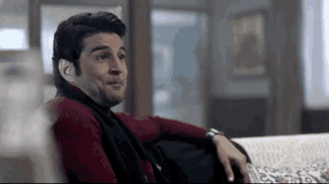 a man in a red sweater and black scarf is sitting on a couch and making a funny face .