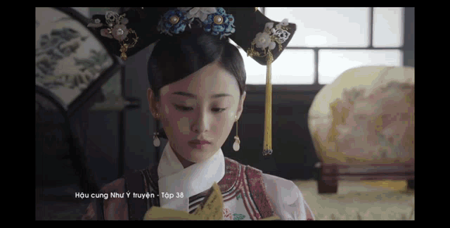 a woman in a traditional costume is sitting in a room with the number 38 on the bottom of the screen