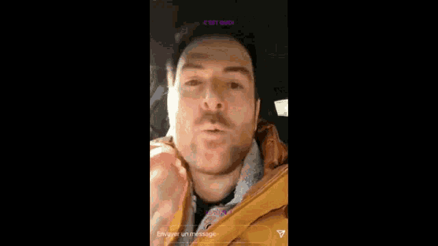 a man in a yellow jacket is making a funny face while sitting in a car with his mouth open .