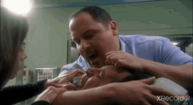 a man in a blue shirt is holding a man 's face in a hospital room with xrecorder in the corner