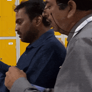 a man in a blue shirt is talking to another man in front of yellow lockers with the number 12 on them