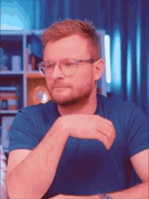 a man wearing glasses and a blue shirt is sitting at a table with his hand on his chin .