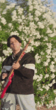 a man is holding a guitar in front of a bush with white flowers