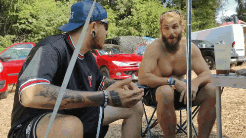 a shirtless man sits at a table talking to a shirtless man