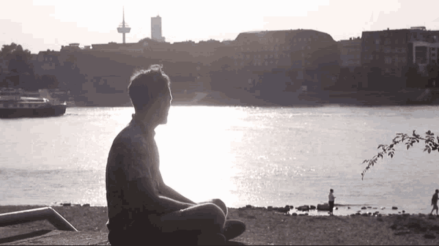 a man sits on the shore of a lake with a boat in the background