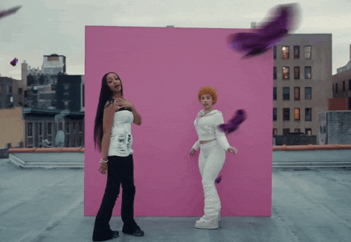 two women standing in front of a pink wall with purple feathers flying around them