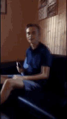 a man in a blue shirt is sitting on a black couch holding a cell phone