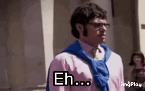 a man in a pink shirt and blue sweater is standing in front of a building and saying eh .