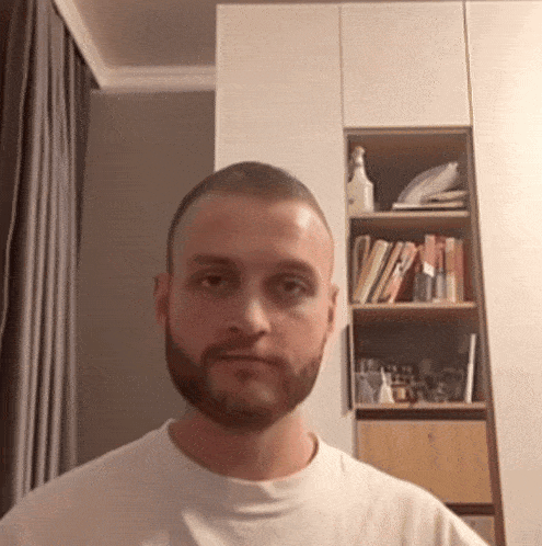 a man with a beard wearing a white shirt is standing in front of a bookshelf