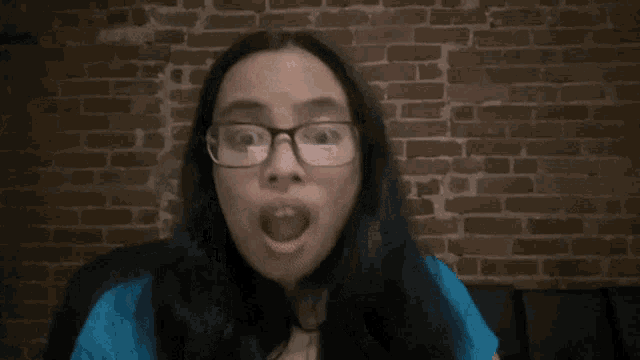 a woman with glasses covering her mouth with her hands in front of a brick wall