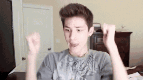 a young man in a gray shirt is making a funny face while raising his arms in the air .