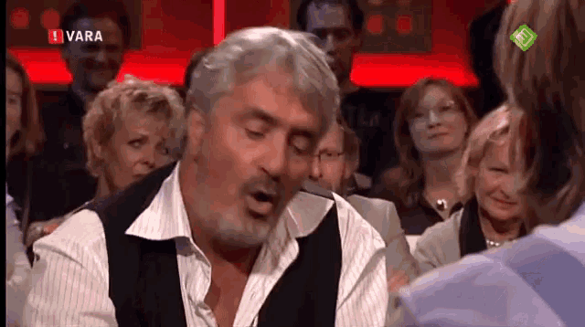 a man with gray hair and a beard talks to a woman in front of a screen that says ' vara ' on it