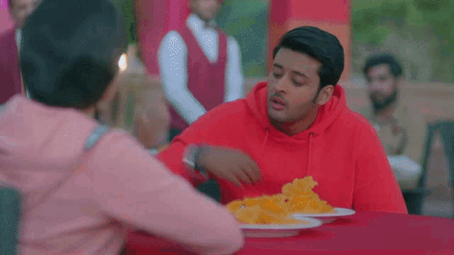 a man and a woman are sitting at a table with plates of food . the man is wearing a red hoodie .