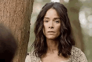 a woman is standing next to a tree in the woods looking at the camera .