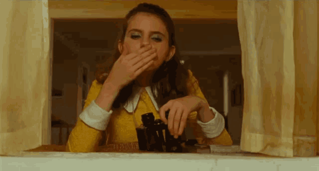 a woman in a yellow dress is looking through binoculars while covering her mouth .