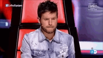 a man in a denim shirt is sitting in a chair with his hands folded .