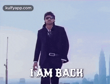 a man in a suit and sunglasses is standing in front of a city with the words `` i am back '' written on it .