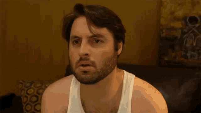 a man with a beard wearing a white tank top is sitting on a couch