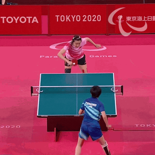 a woman in a pink shirt is playing ping pong in front of a sign that says toyota