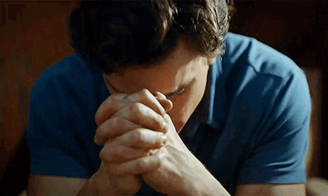 a man in a blue shirt is praying with his hands folded in front of his face .
