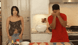 a man in a red shirt is covering his face with his hands while a woman stands behind him