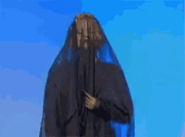 a woman in a black dress with a blue veil on her head is standing in front of a blue sky .
