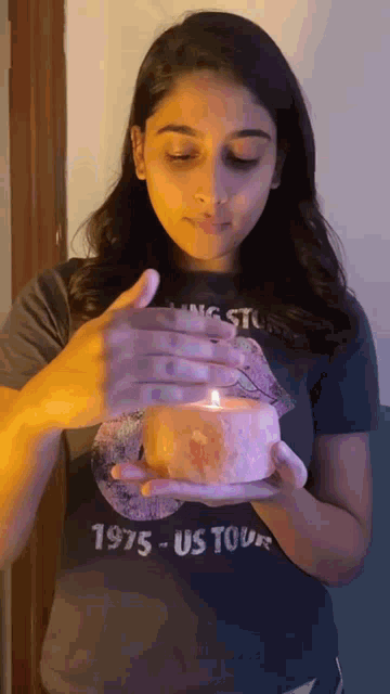 a woman in a 1975 us tour shirt is holding a lit candle .