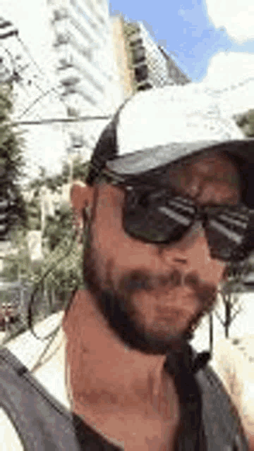 a man with a beard and sunglasses is wearing a hat and a backpack .