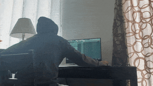 a man in a hoodie sits at a desk using a computer