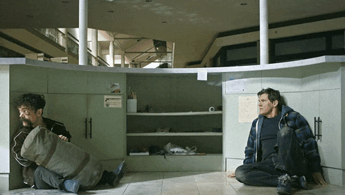 two men are sitting on the floor in an empty room with a sign on the wall that says ' emergency '