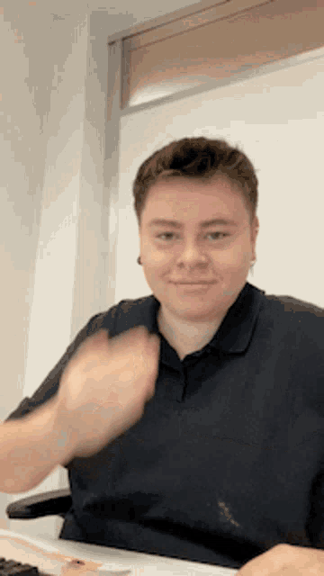 a man in a black shirt is sitting at a desk and waving his hand