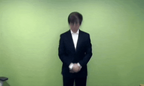 a man in a suit is standing in front of a green wall with his hands folded .