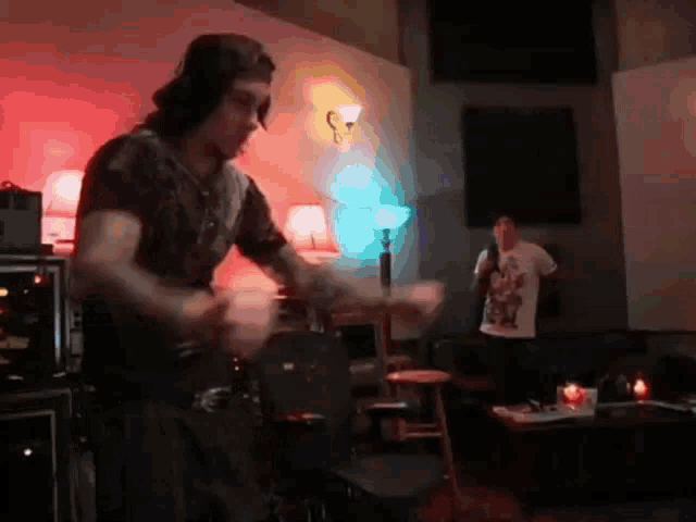 a man in a baseball cap is playing drums in a room while another man looks on .