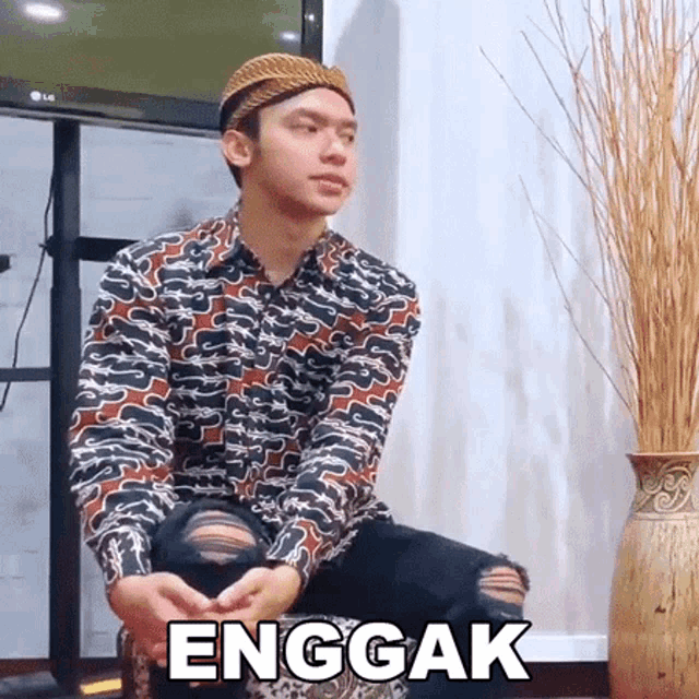 a man wearing a hat and a floral shirt is sitting in front of a vase with the word enggak on it .