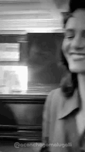a black and white photo of a woman smiling while standing in front of a refrigerator .