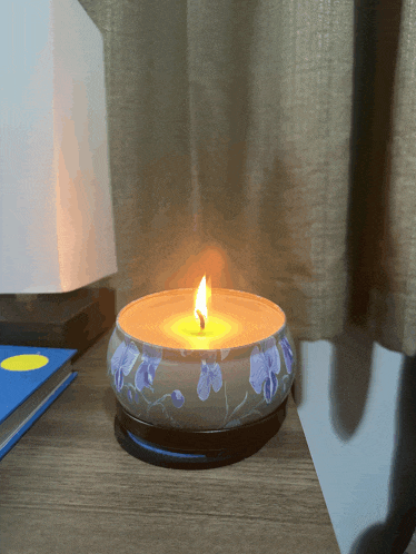 a candle with purple flowers on it sits on a table next to a lamp