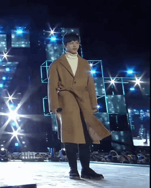a man wearing a brown coat and a white turtleneck is standing on a stage
