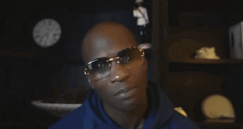 a bald man wearing sunglasses is looking at the camera with a clock in the background