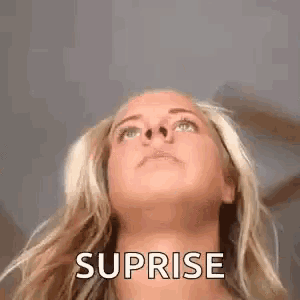 a woman with blonde hair is looking up at the sky with the word suprise written on her face .
